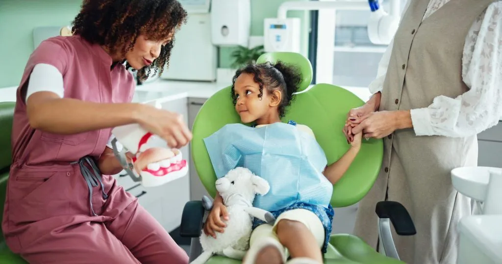 A friendly female dental professional demonstrates brushing techniques using a large model of teeth to reassure a young girl sitting in a green dental chair.