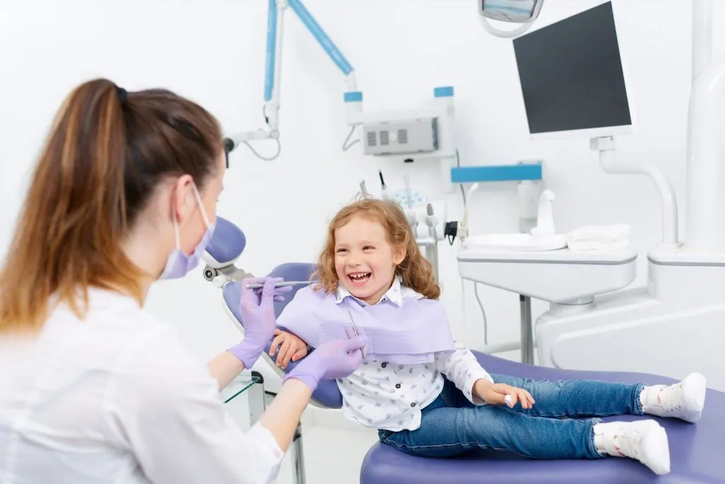 A smiling young girl sits in a pediatric dental chair while a female dentist examines her teeth.