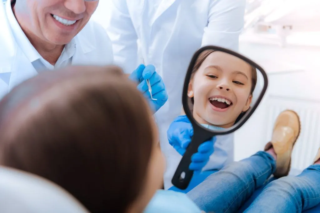 A smiling young girl looks into a hand mirror during a routine checkup at a pediatric dental clinic.