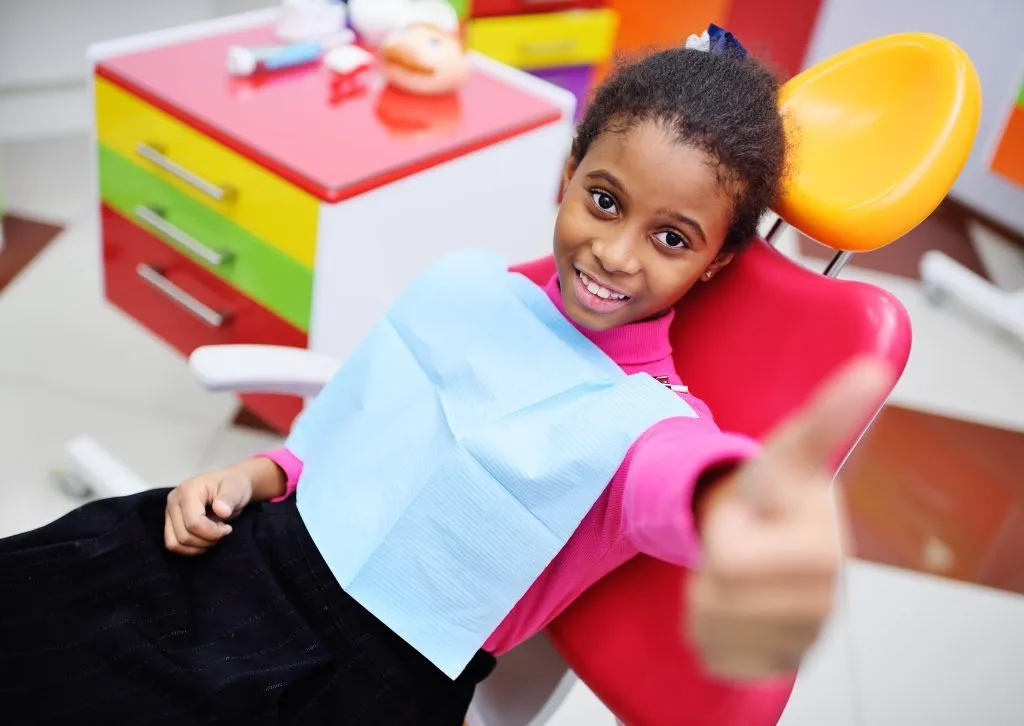 A young girl giving a thumbs-up while sitting in a pediatric dental chair.