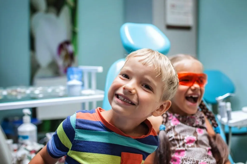 A happy young boy and girl smiling cheerfully while sitting inside a pediatric dental office.