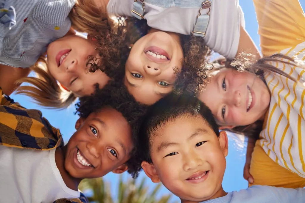 A low-angle close-up photograph of a diverse group of young children looking down and smiling into the camera.