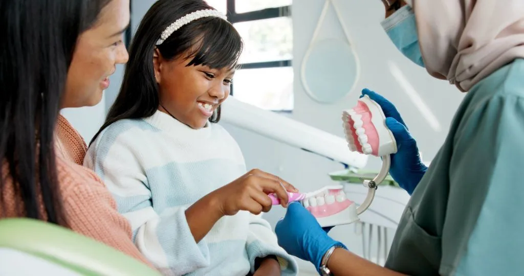 A dentist uses a large anatomical model of teeth to show a smiling young girl and her mother how to brush properly.